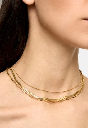 Zbliżenie na szyję kobiety noszącej trzy złote naszyjniki warstwowe o różnych stylach łańcuchów na jasnej skórze.