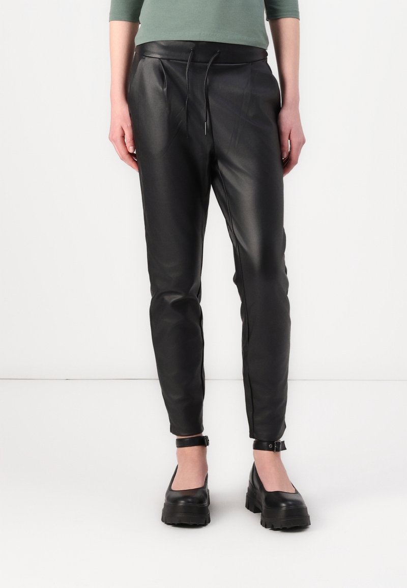 Personne portant un pantalon de jogging en cuir noir avec cordon à la taille et des chaussures à plateforme noires avec bride à la cheville, debout sur un fond blanc.