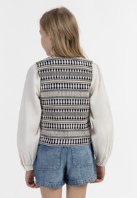 Sweter bez rękawów z wzorzystym deseniem w kolorze granatowym, kremowym i złotymi akcentami, połączony z białą, fakturowaną bluzką z długim rękawem i dżinsowymi szortami.