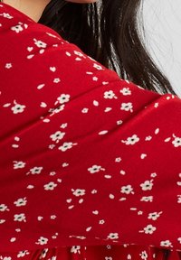 Femme portant un tissu rouge avec un petit motif floral blanc et ayant les cheveux raides et foncés, visible sur un fond gris.