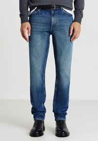 Uomo che indossa jeans blu, cintura nera, maglione grigio e stivali in pelle nera, in piedi su uno sfondo bianco uniforme.