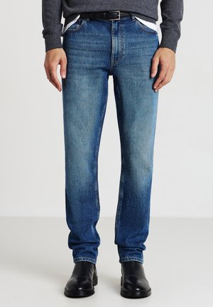 Mann in blauen Jeans, schwarzem Gürtel, grauem Pullover und schwarzen Lederstiefeln steht vor einem schlichten weißen Hintergrund.
