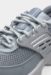 Κοντινή λήψη ανοιχτογκρί αθλητικού παπουτσιού με υφασμάτινο πλέγμα, ανακλαστικά συνθετικά επάνω στρώματα και χοντρές λευκές και γκρί κορδόνια με σχέδιο, περασμένες μέσα από μεταλλικά οπές.