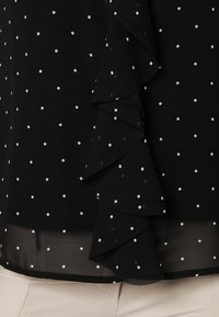 Blusa negra con lunares blancos, que presenta un detalle de volantes en la parte delantera. Hecha de una tela ligera, con un dobladillo transparente.