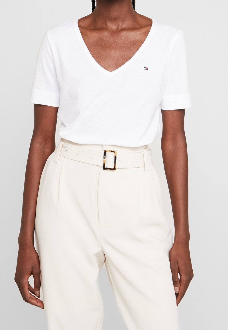 Biała bawełniana koszulka z dekoltem w serek i krótkimi rękawami, z małym czerwono-niebieskim logo na piersi. Zestawiona z jasnokremowymi spodniami na wysokim stanie.