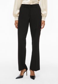 Kvinne iført svarte skreddersydde slengbukser, kremfarget bluse med smockede mansjetter, og svarte høyhælte spisse sko, står foran en hvit bakgrunn.