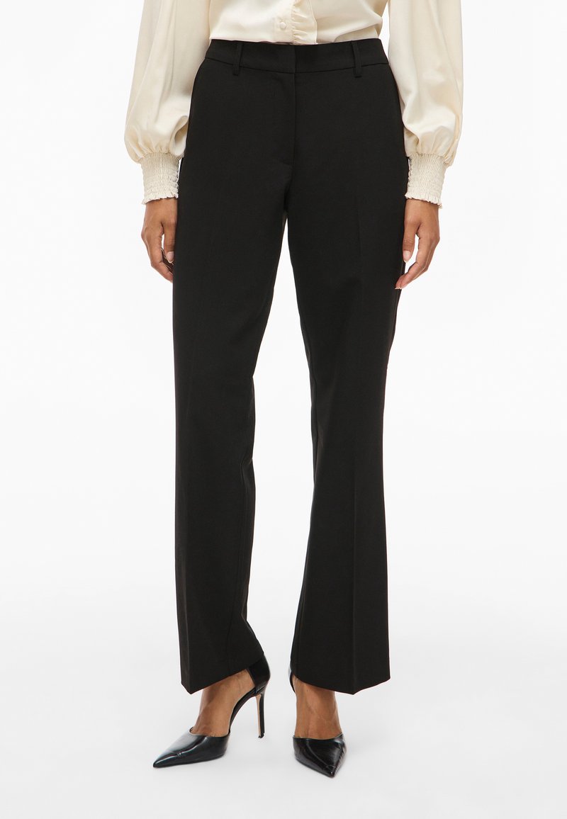 Kvinne iført svarte skreddersydde slengbukser, kremfarget bluse med smockede mansjetter, og svarte høyhælte spisse sko, står foran en hvit bakgrunn.