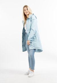 Femeie blonda îmbrăcată cu o jachetă impermeabilă cu glugă de culoare albastru deschis, cămașă albă, blugi și adidași albi, stând în fața unui fundal alb.