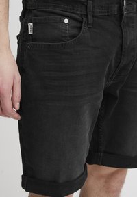 Shorts din denim negru cu tiv rulat, design cu cinci buzunare și accente din metal. Textură netedă și aspect ajustat evidențiate.