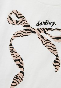 Biały materiał z haftowanym wzorem różowej i czarnej wstążki w paski zebry. Słowo "darling" jest wyszyte na czarno nad wstążką.