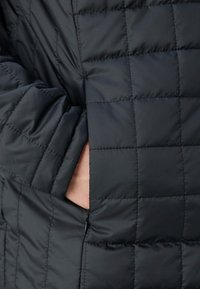 Main partiellement insérée dans la poche latérale d'une veste matelassée noire avec un motif de piqûres carrées.