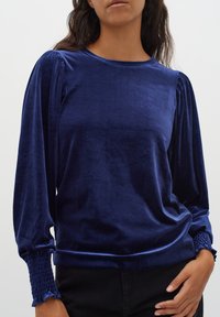Marinblå långärmad topp i sammet med rund halsringning, volangärmar och smockade ärmslut, vilket ger en strukturerad look.