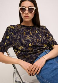 Marineblå kortærmet top med et geometrisk mønster i guld, parret med lyseblå denim. Model iført hvide katteøjne solbriller, siddende.