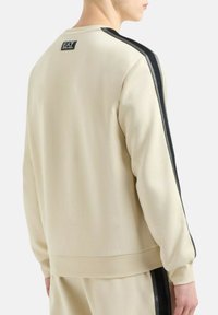 Persona che indossa una felpa beige con strisce nere sulle spalle e pantaloni beige abbinati, mostrata di spalle e di lato.