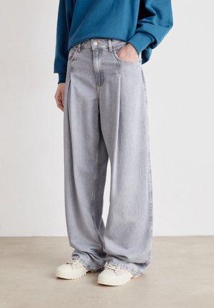 Persoon die lichtgrijze jeans met wijde pijpen, witte sneakers en een blauw shirt met lange mouwen draagt, staat met één hand in de zak.