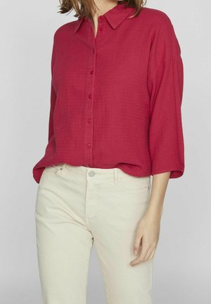 Kobieta ubrana w luźną, czerwoną koszulę na guziki z długimi rękawami, włożoną do kremowych spodni z wysokim stanem.