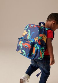 Mladé dieťa so štýlovým batohom s farebným vzorom dinosaurov, v červenom tričku, tmavých džínsoch a bielych teniskách, stojace pred neutrálnym pozadím.