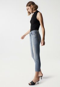 Top negro ajustado sin mangas combinado con jeans de tiro alto desgastados de color azul claro y sandalias de tacón negras. Enfocarse en los detalles de la forma y el ajuste.