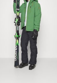 Persone che indossa una giacca verde, pantaloni da sci neri, guanti e stivali, mentre tiene verticalmente degli sci Salomon neri e verdi.