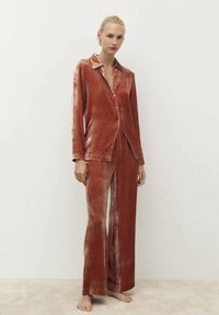 Completo in velluto a due pezzi di colore ruggine, composto da una camicia a maniche lunghe con colletto e bottoni, e pantaloni a gamba larga, con una texture morbida e una vestibilità comoda.