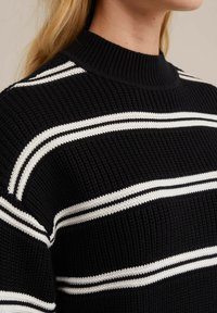 Svart stickad tröja med vita horisontella ränder. Har en ribbad halsringning och strukturerad tyg. Närbild av axeln och halsringningsområdet.