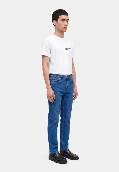 Jeune homme debout portant un T-shirt blanc à manches courtes avec un petit texte, un jean bleu et des mocassins noirs sur un fond clair uni.
