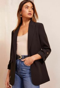 Schwarzer Blazer mit schmaler Passform und Revers, mit hochgekrempelten Ärmeln und einem einzelnen Knopfdetail, getragen über einem beigen Tanktop.