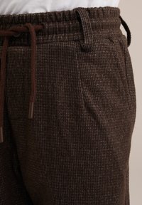 Bruna strukturerade byxor med dragsko i midjan, sidofickor och ett subtilt rutmönster, tillverkade av mjukt, vävt tyg.