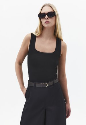 Kvinde med langt blondt hår iført sort ærmeløs top, sorte bukser med vide ben, sort bælte og mørke ovale solbriller stående mod en hvid baggrund.