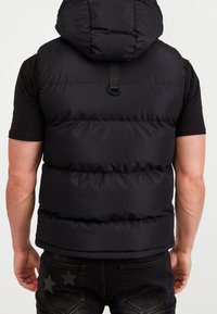 Uomo che indossa un gilet puffer nero con cappuccio sopra una maglietta nera a maniche corte e jeans neri con toppe a forma di stella sulle tasche posteriori, visto da dietro.