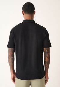 Chemise à manches courtes en tissu noir texturé avec col à boutons. Coupe droite avec fines rayures verticales et coupe décontractée.