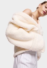 Hedvábný krémový oversized kabát s plyšovou texturou, ohrnutými rukávy a šálovým límcem, kombinovaný se světlými pruhovanými kalhotami.