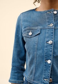 Nainen, jolla on päällään sininen farkkutakki, jossa on hopeiset napit, kolmannes hihasta ja kaulukseton pääntie vaaleaa taustaa vasten.