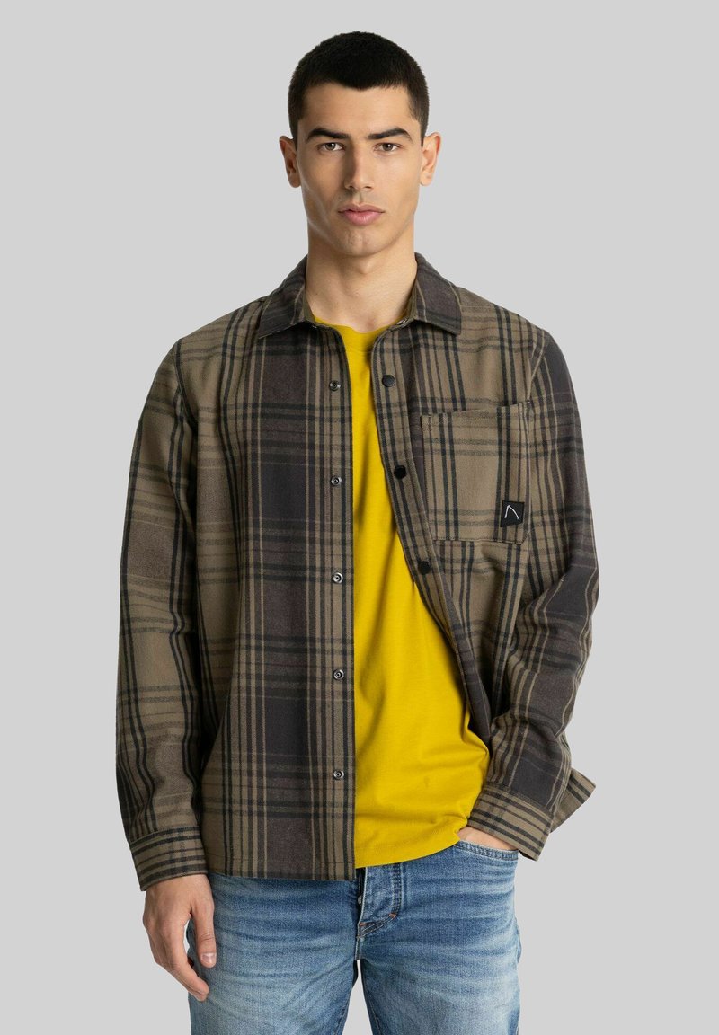 Jongeman met kort haar, gekleed in een bruin en zwart geruite overhemdo over een geel T-shirt en blauwe jeans, staande tegen een grijze achtergrond.