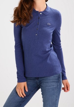 Kobieta ubrana w dopasowaną, długorękawową niebieską koszulkę polo z guzikami i dżinsy, pozująca z jedną ręką na udzie na białym tle.