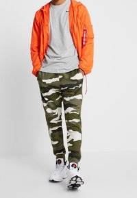 Person klädd i en orange jacka, grå t-shirt, grön och vit kamouflage Nike joggers, och vita sneakers med en färgglad logotyp, med händerna i fickorna.