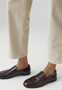 Scarpe slip-on bordeaux con una finitura in pelle lucida, caratterizzate da dettagli cuciti e una suola nera bassa, abbinate a pantaloni in corduroy beige.