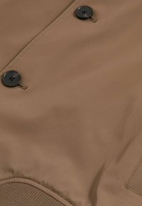 Brunt stof med en glat tekstur, der har to sorte knapper og et kontrasterende ribbet afsnit. Designet er struktureret og minimalisk.