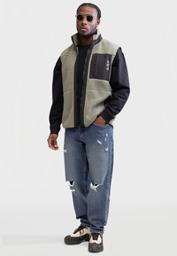 Alyvuogių žalumo fliiso liemenė su juodu kišeniu, dėvima ant juodos palaidinės. Mėlyni, vėrimo stiliaus džinsai ir smėlio spalvos sportbačiai užbaigia aprangą.