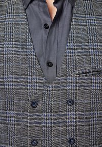 Kamizelka w szaro-niebieski wzór w kratę z dekoltem w serek, mająca podwójne zapięcie i ciemne guziki; noszona na gładkiej szarej koszuli.