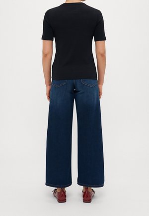 Person vendt væk iført en sort kortærmet skjorte, vidde ben blå jeans og skinnende røde snøresko mod en hvid baggrund.