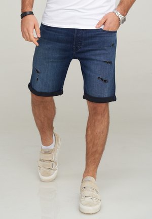 Shorts de mezclilla en azul oscuro con dobladillos enrollados y detalles desgastados. Combinados con zapatos beige con correas ajustables.