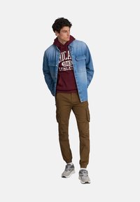 Jeansowa kurtka na bordo bluzie z kapturem, w zestawieniu z brązowymi spodniami cargo i szarymi sneakersami. Kurtka ma zapięcia na guziki i kieszeń na piersi.