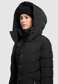 Crna puhača jakna s velikim kapuljačom, sa šivanim uzorkom i bočnim džepovima. Ima zatvaranje na zip i brendirani detalj na rukavu.