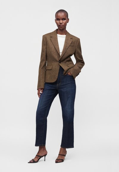 Femme portant un blazer marron sur un top blanc, un jean droit bleu foncé et des talons ouverts noirs sur un fond blanc uni.
