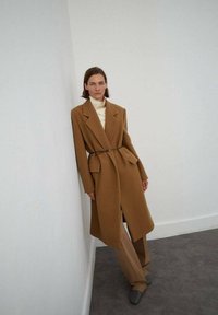 Lungo cappotto sartoriale color cammello, con vita regolabile, grandi tasche e revers accentuati, abbinato a un dolcevita crema e pantaloni beige.