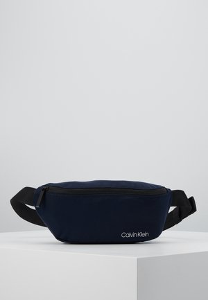 Granatowa nerka Calvin Klein z czarnym regulowanym paskiem i zamkiem błyskawicznym, umieszczona na białej powierzchni na jednolitym szarym tle.