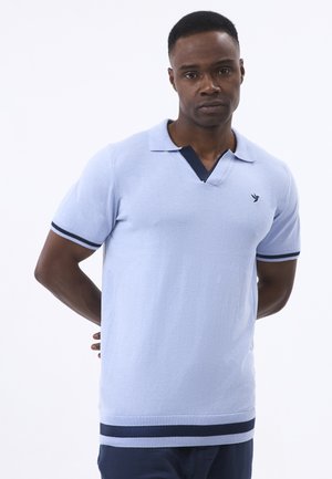 Férfi világoskék rövid ujjú galléros ingben, sötétkék díszítéssel és kis madár logóval, kezét a hátán tartva, fehér háttér előtt áll.