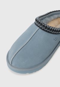 Pantofole in suede blu con foderatura in pelliccia crema, cuciture decorative nere lungo il colletto e suola in gomma. Logo del marchio visibile sul lato.