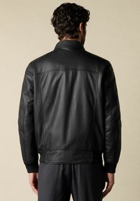 Man met donker krullend haar draagt een zwart leren jack en donkere broek, staat met zijn rug naar de camera tegen een effen achtergrond.